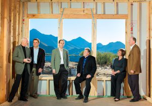 Six executives standing in window of framed-in house with mountains in background.
